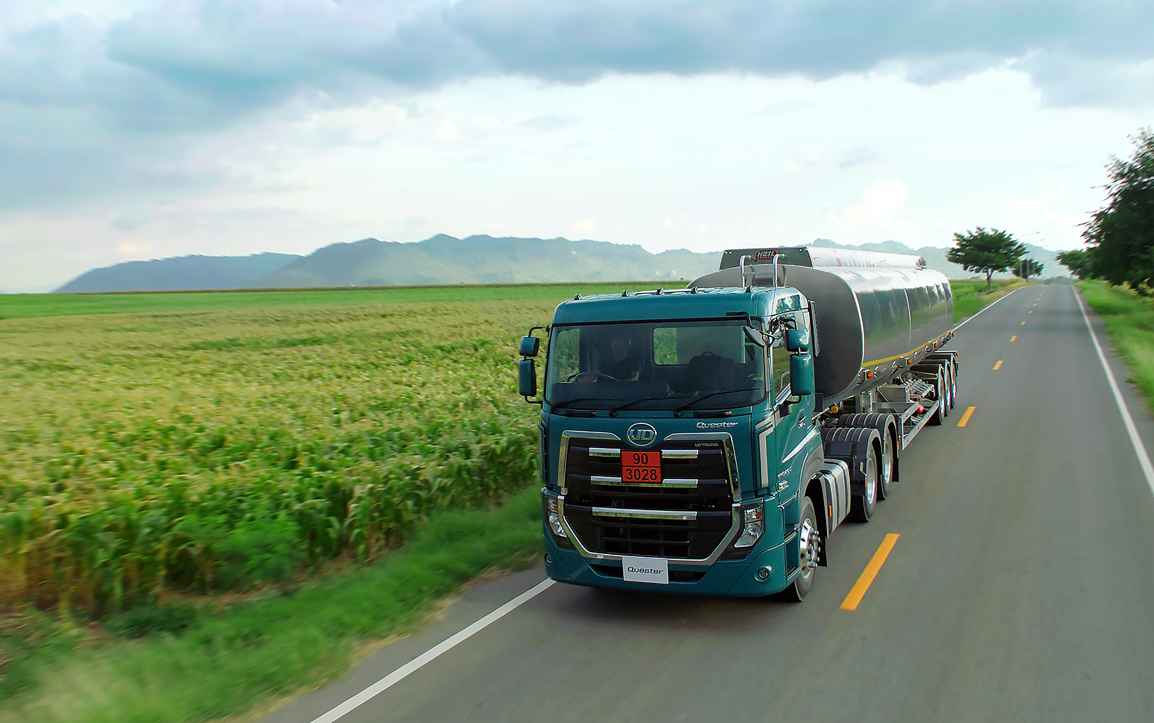 Quester Tanker truck driving on rural road between green fields under a cloudy sky.