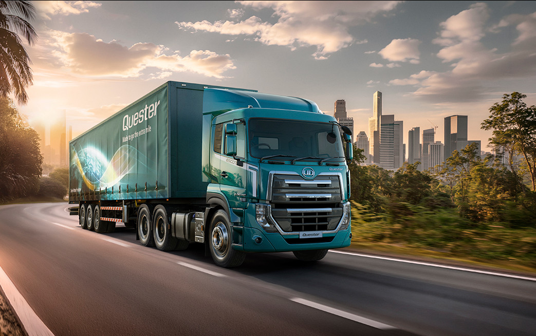 Blue semi-truck driving on a highway with a city skyline in the background at sunset.