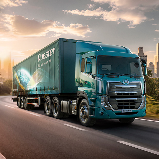 Blue semi-truck driving on a sunny road with a city skyline in the background.