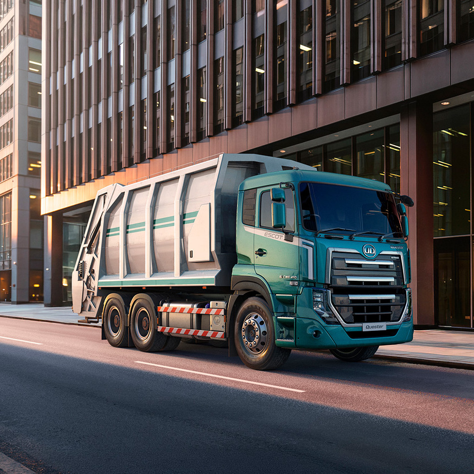 Green garbage truck driving down an empty city street surrounded by tall buildings.