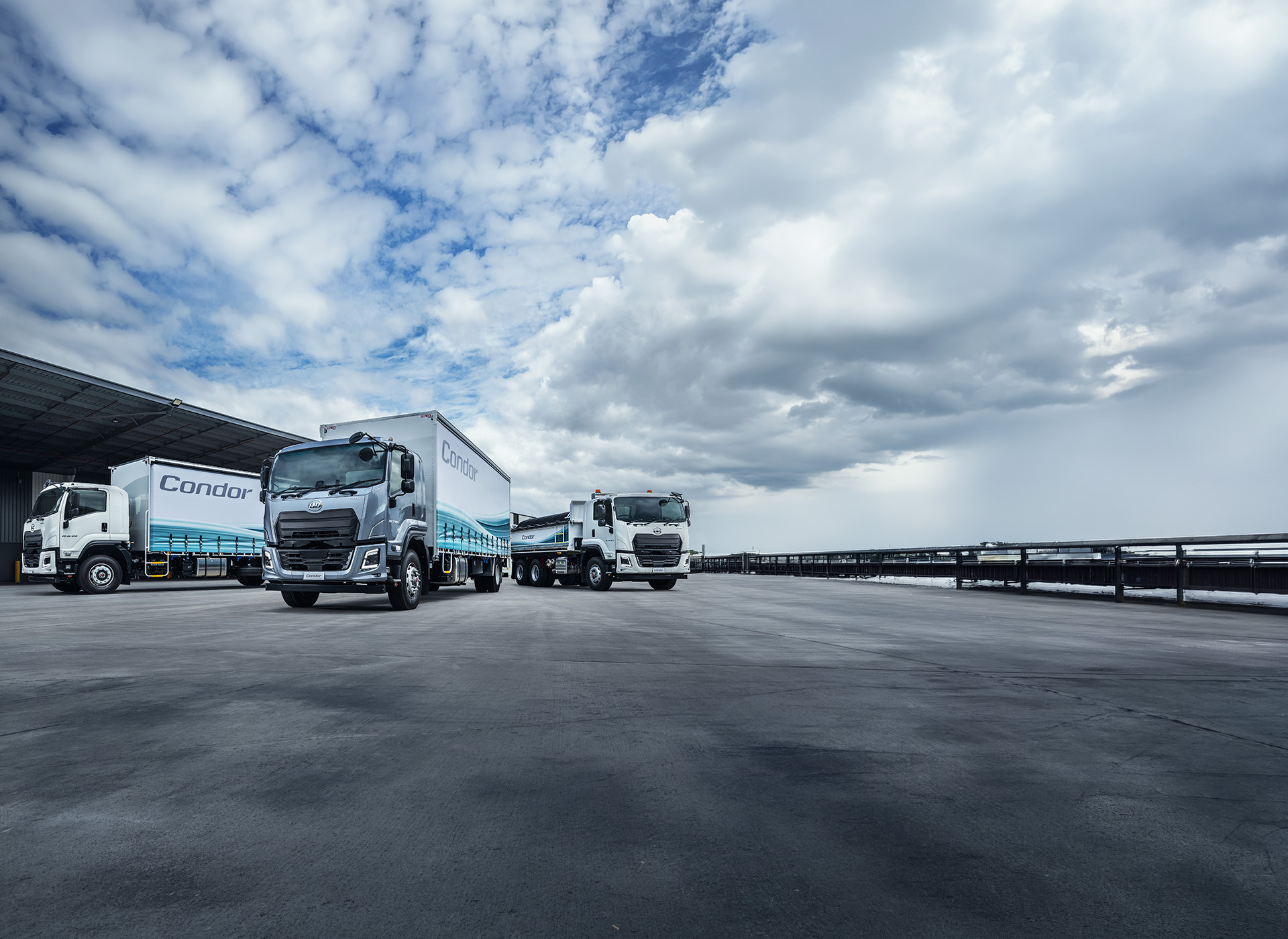 Three delivery trucks parked near a warehouse under a cloudy sky.