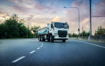 White semi-truck driving on a curved road at sunset with trees in the background.