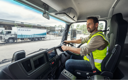 Truck driver in high-visibility vest sits at the wheel, warehouse and truck visible outside.