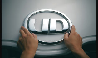 Hands holding a silver UD logo emblem on a gray surface.