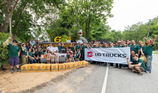 กลุ่มคนยืนถ่ายรูปกลางแจ้งถือป้าย UD TRUCKS ท่ามกลางต้นไม้และธรรมชาติ