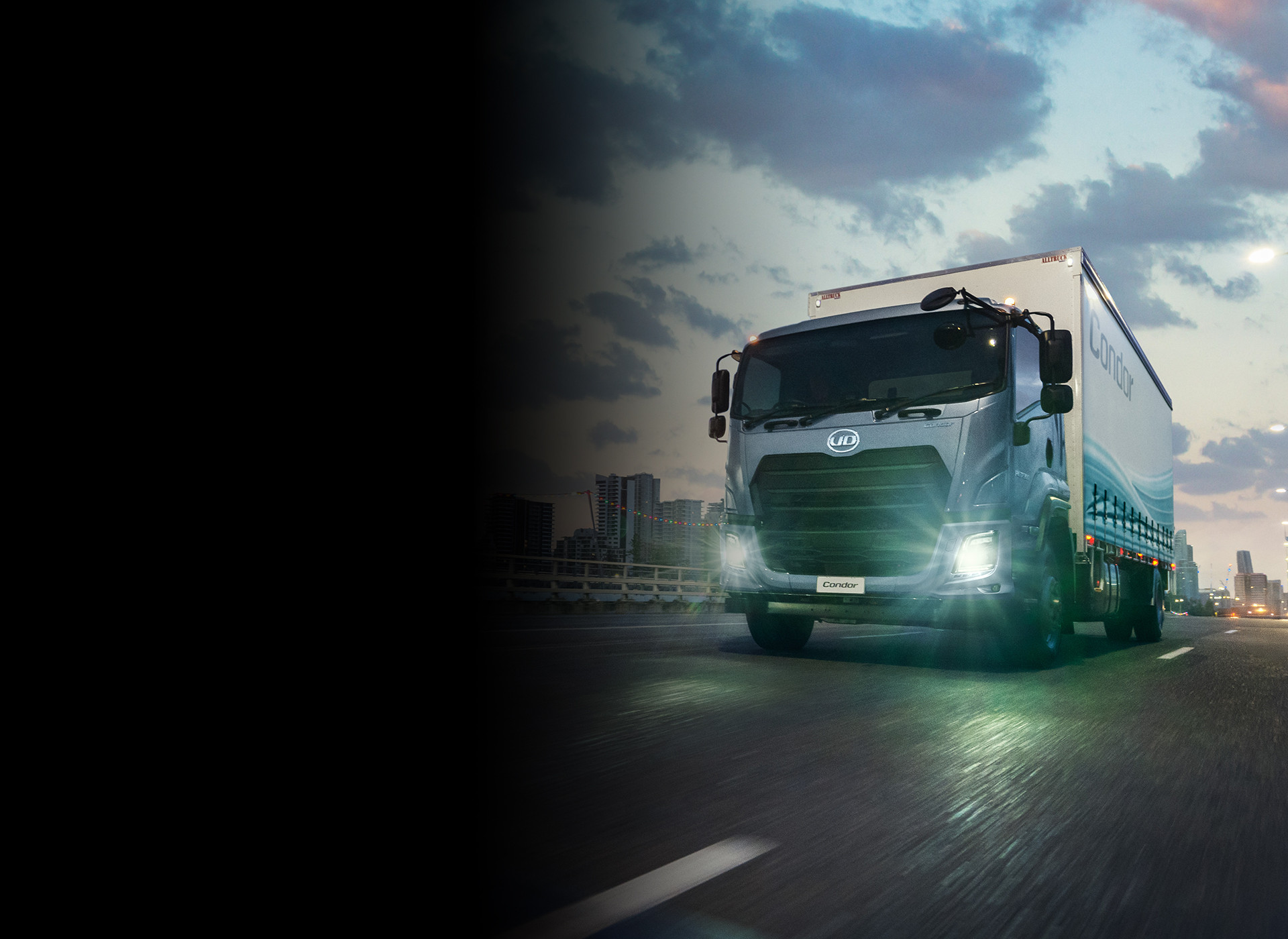 Large truck driving on a city road at dusk with headlights on.