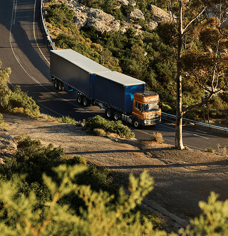 Xe tải lớn màu cam và xanh di chuyển trên đường quanh co giữa rừng cây.