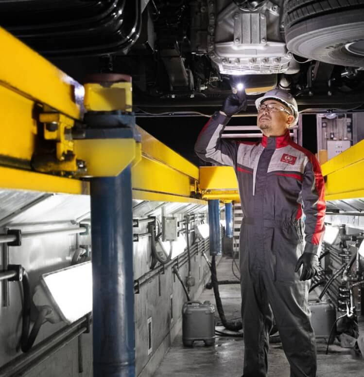 Mechanic in uniform inspecting a vehicle from below with a flashlight in a well-lit garage.