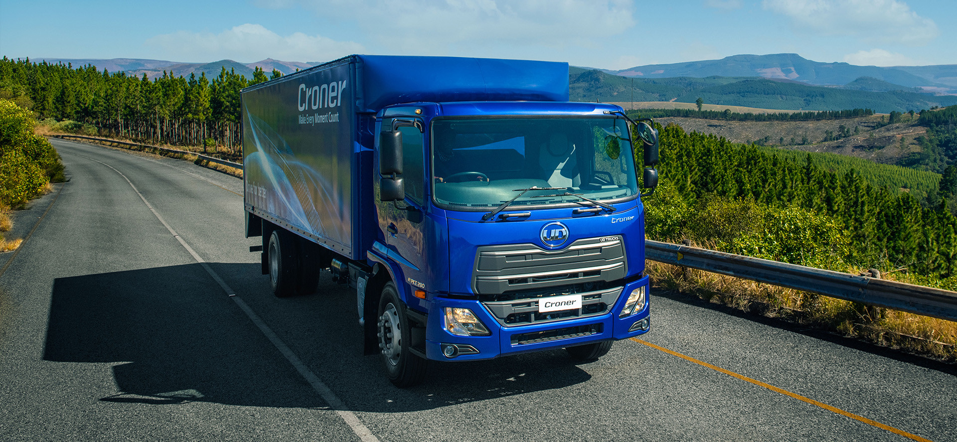 Blue delivery truck driving on a road, surrounded by green hills and trees.
